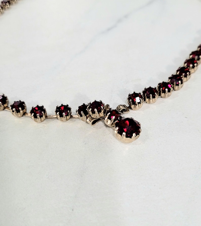May include: A silver-toned necklace adorned with a sequence of round, deep red gemstones. The necklace showcases a classic design, with a single, larger gemstone pendant. The gemstones are evenly spaced along the chain.