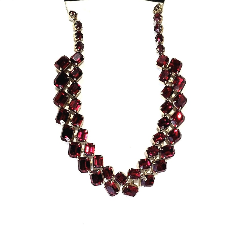 May include: A striking necklace with a series of rectangular, deep red gemstones set in silver-toned metal. The stones are arranged in a cascading pattern, creating a bold design. The necklace is displayed against a white background.