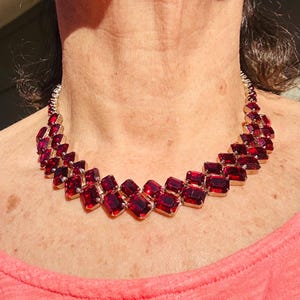 May include: A gold-coloured necklace with a series of rectangular, deep red gemstones. The stones are set closely together, creating a bold design. The necklace is a statement piece, ideal for adding a touch of glamour to an outfit.