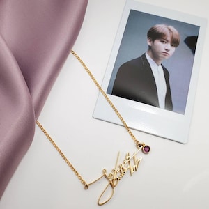 A gold-colored chain necklace with a cursive signature pendant and a pink gemstone rests next to a polaroid photo of a man and a lavender fabric fold.