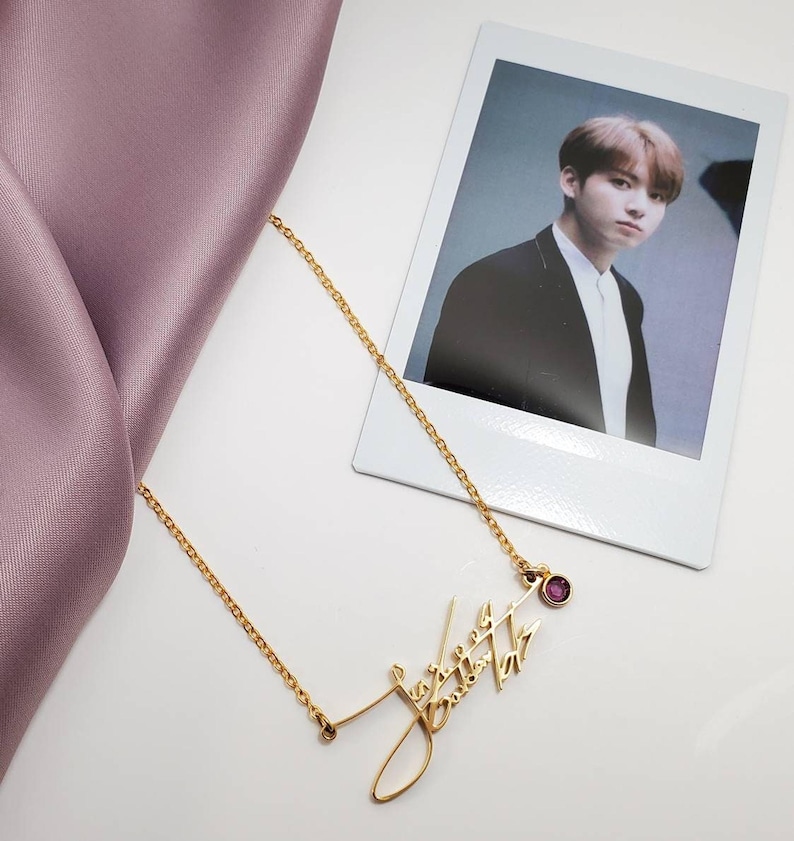 A gold-colored chain necklace with a cursive signature pendant and a pink gemstone rests next to a polaroid photo of a man and a lavender fabric fold.
