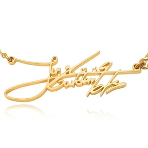 A gold-colored chain necklace features a cursive signature pendant and a small circular pink gemstone charm.
