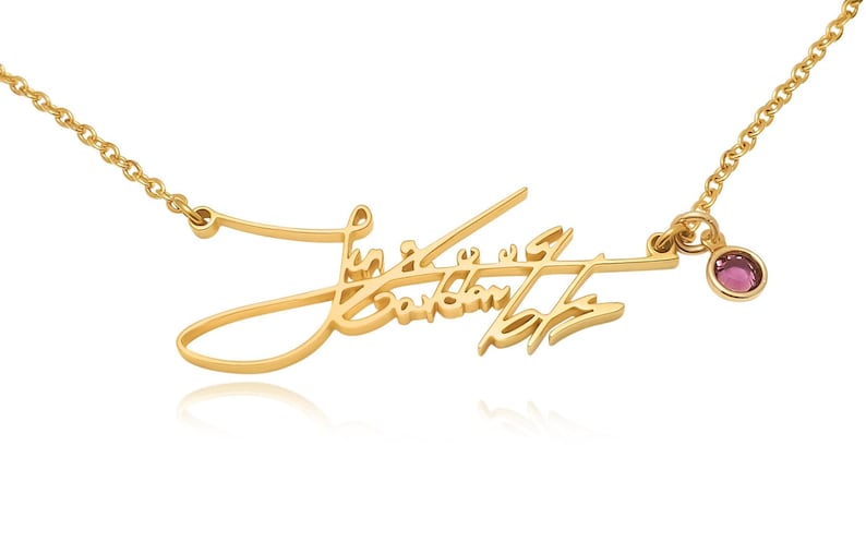 A gold-colored chain necklace features a cursive signature pendant and a small circular pink gemstone charm.