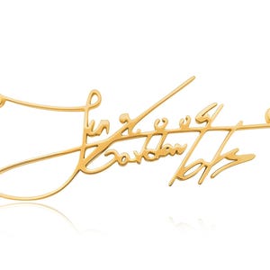 Close-up of a gold-colored chain necklace with a cursive signature pendant against a white background.