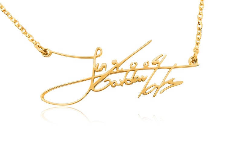 Close-up of a gold-colored chain necklace with a cursive signature pendant against a white background.