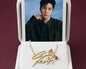 May include: A gold-toned necklace featuring a stylized signature pendant and a small round red gemstone charm, displayed in a white velvet-lined box alongside a rectangular photograph of a person wearing a black jacket against a blue background.