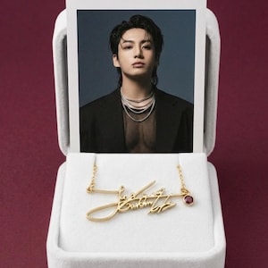 A gold-colored metal signature pendant necklace and a polaroid photo of a man in a black jacket are displayed inside an open white velvet box on a dark red background.