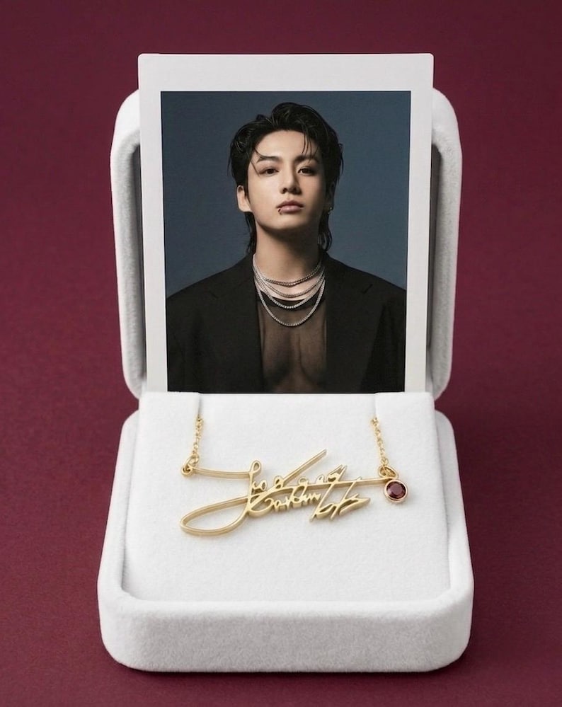 A gold-colored metal signature pendant necklace and a polaroid photo of a man in a black jacket are displayed inside an open white velvet box on a dark red background.