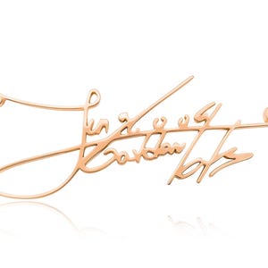 Close-up of a rose-gold-colored chain necklace with a cursive signature pendant against a white background.