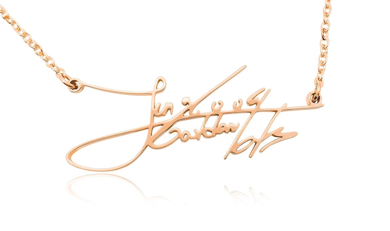 Close-up of a rose-gold-colored chain necklace with a cursive signature pendant against a white background.