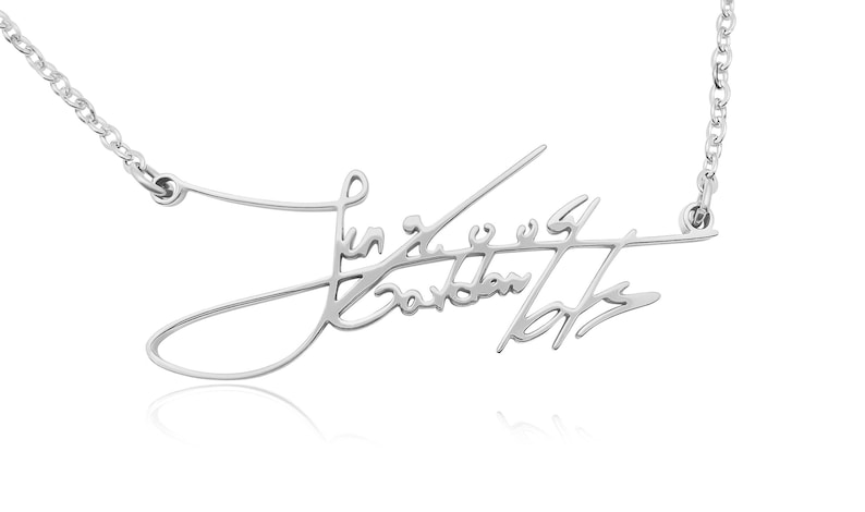 Close-up of a silver-colored chain necklace with a cursive signature pendant against a white background.
