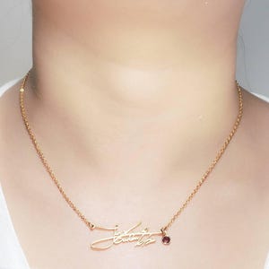A person wearing a gold-colored chain necklace with a cursive signature pendant and a small pink gemstone charm.