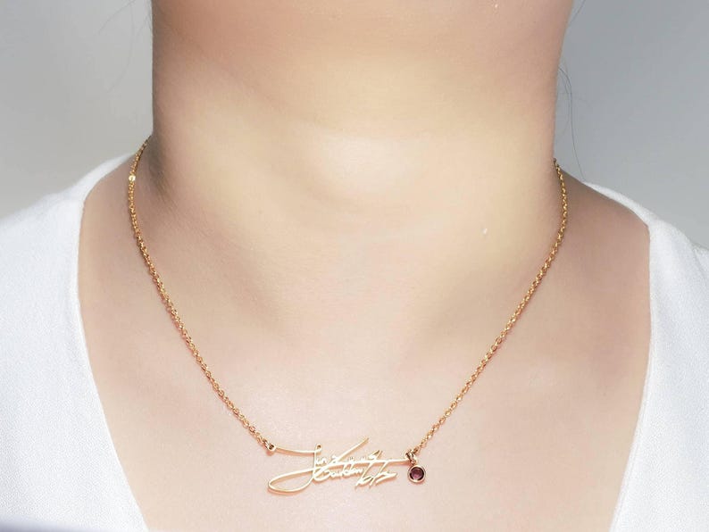 A person wearing a gold-colored chain necklace with a cursive signature pendant and a small pink gemstone charm.