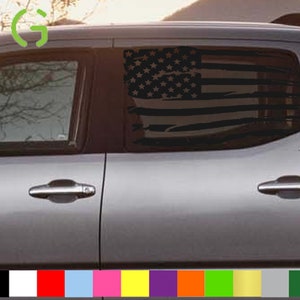 Puede incluir: Una camioneta plateada con una calcomanía de bandera estadounidense en blanco y negro en la ventana trasera. La calcomanía es un diseño desgastado con un aspecto descolorido. La camioneta tiene una manija de la puerta negra y un parachoques plateado.