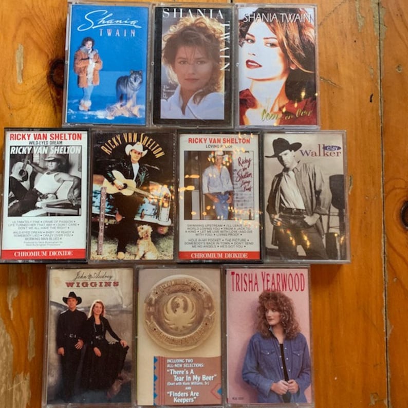 May include: A collection of eight cassette tapes featuring country music artists Shania Twain, Ricky Van Shelton, Clay Walker, John M. Wiggins, and Trisha Yearwood. The tapes are arranged in two rows on a wooden surface.