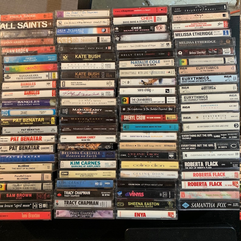 May include: A collection of vintage cassette tapes featuring various artists, including All Saints, Cher, Eurythmics, Pat Benatar, and more. The tapes are arranged in rows on a shelf, with the titles and artist names visible.