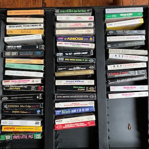 May include: A close-up view of a black cassette tape storage unit with a variety of cassette tapes. The tapes are organized by artist and album title. Some of the artists include Bryan Adams, INXS, The Neville Brothers, Robert Plant, Maxi Priest, Bonnie Raitt, R-S-F, Tone-Loc, Wilson Phillips, Enigma, Martika, MCJ & Cool G, Blue Sky, Mali Van, Mister Mixi & Skinny Scotty, Extreme, Green Jelly, and Three Little Pigs.