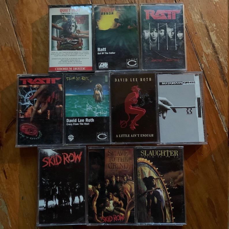 May include: A collection of vintage cassette tapes featuring album art from various rock and heavy metal bands. Titles include "Quiet Riot," "Ratt," "David Lee Roth," "Skid Row," and "Slaughter." The tapes are arranged on a wooden surface.