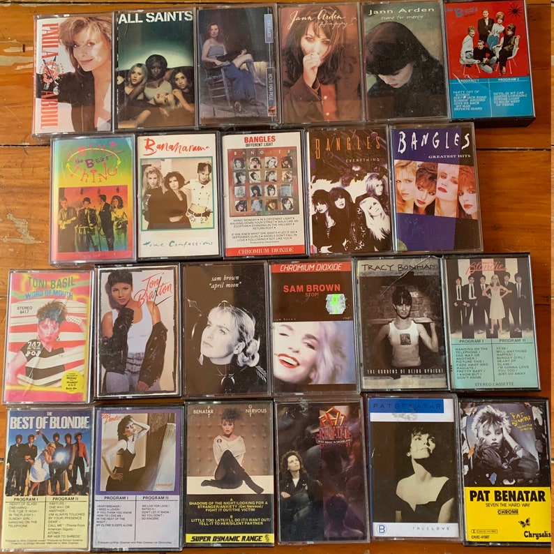 May include: A collection of cassette tapes featuring various artists, including Paula Abdul, All Saints, Jann Arden, Bananaram, The Bangles, Toni Basil, Sam Brown, Tracy Bonham, Pat Benatar, and Blondie. The tapes are arranged in rows on a wooden surface.
