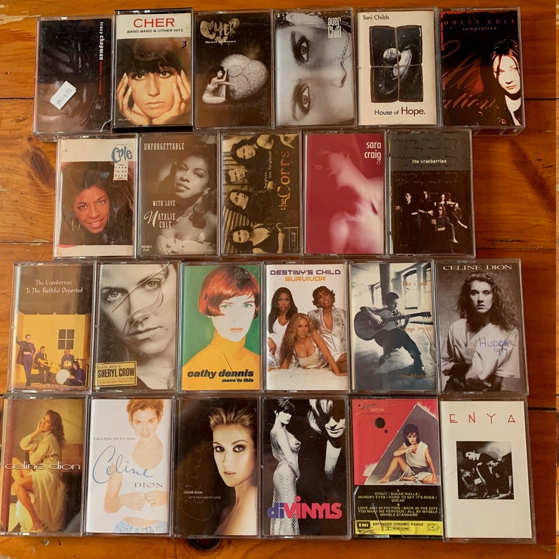 May include: A collection of vintage cassette tapes featuring various artists, including Cher, Celine Dion, The Cranberries, and more. The tapes are arranged in rows on a wooden surface.