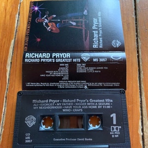 May include: A black cassette tape with a white label. The label has a picture of comedian Richard Pryor and the text "Richard Pryor - Richard Pryor's Greatest Hits". The text on the label lists the songs on the cassette tape: "Ali", "Exorcist", "My Father", "Nigger with a Seizure", "My Neighborhood", "Have Your Ass Home by 11:00", "Wino", "Craps". The cassette tape has a black case with a white label on the front and a black label on the back. The back label has the text "Richard Pryor", "Richard Pryor's Greatest Hits", "Side One", "Side Two", and a list of songs on each side. The back label also has the text "Executive Producer David Banks", "1977 Warner Bros. Records Inc.", "Unauthorized reproduction of this recording is prohibited by law and subject to criminal prosecution", "Distributed by/Distribu&eacute; Par WEA Music of Canada Ltd/WEA Musique Du Canada Ltee 1810 Birchmount Rd., Scarborough, Ontario", "A Warner Communications Company".