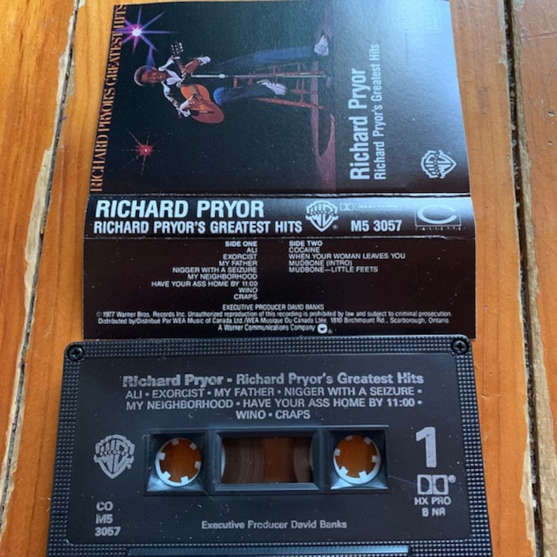 May include: A black cassette tape with a white label. The label has a picture of comedian Richard Pryor and the text "Richard Pryor - Richard Pryor's Greatest Hits". The text on the label lists the songs on the cassette tape: "Ali", "Exorcist", "My Father", "Nigger with a Seizure", "My Neighborhood", "Have Your Ass Home by 11:00", "Wino", "Craps". The cassette tape has a black case with a white label on the front and a black label on the back. The back label has the text "Richard Pryor", "Richard Pryor's Greatest Hits", "Side One", "Side Two", and a list of songs on each side. The back label also has the text "Executive Producer David Banks", "1977 Warner Bros. Records Inc.", "Unauthorized reproduction of this recording is prohibited by law and subject to criminal prosecution", "Distributed by/Distribu&eacute; Par WEA Music of Canada Ltd/WEA Musique Du Canada Ltee 1810 Birchmount Rd., Scarborough, Ontario", "A Warner Communications Company".
