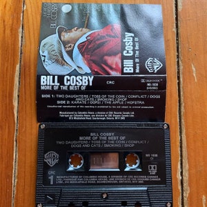 May include: A black cassette tape with a white label. The label has a picture of Bill Cosby and the text "Bill Cosby More of the Best of". The cassette tape is labeled with the text "Bill Cosby More of the Best of" and the track list: "Two Daughters/Toss of the Coin/Conflict/Dogs and Cats/Smoking/Shop".