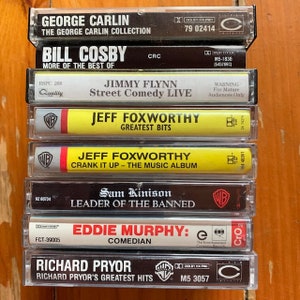 May include: A stack of eight cassette tapes featuring comedy albums by George Carlin, Bill Cosby, Jimmy Flynn, Jeff Foxworthy, Sam Kinison, Eddie Murphy, and Richard Pryor. The tapes are arranged vertically with the titles and artist names visible.