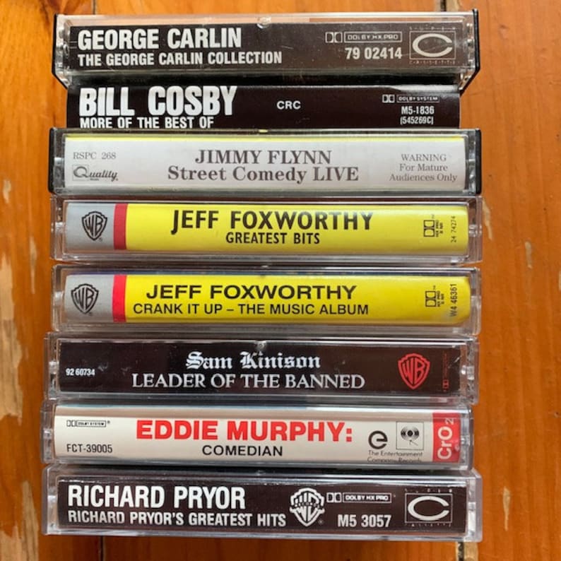 May include: A stack of eight cassette tapes featuring comedy albums by George Carlin, Bill Cosby, Jimmy Flynn, Jeff Foxworthy, Sam Kinison, Eddie Murphy, and Richard Pryor. The tapes are arranged vertically with the titles and artist names visible.