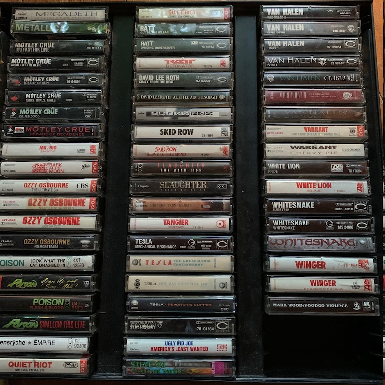 May include: A collection of vintage cassette tapes featuring rock and heavy metal music artists. The tapes are arranged in a black display case, showcasing bands like M&ouml;tley Cr&uuml;e, Ozzy Osbourne, and Van Halen. The labels display album titles and band names.