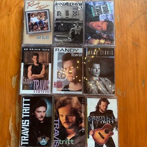 May include: A collection of nine cassette tapes featuring country music artists Randy Travis and Travis Tritt. The tapes are arranged in three rows of three. The top row features Randy Travis albums "Old 8x10", "Storms of Life", and "Randy Travis Greatest Hits Volume One". The middle row features Randy Travis albums "No Holdin' Back", "High Lonesome", and "Randy Travis Greatest Hits Volume One". The bottom row features Travis Tritt albums "Travis Tritt", "It's All About to Change", and "T-R-O-U-B-L-E".