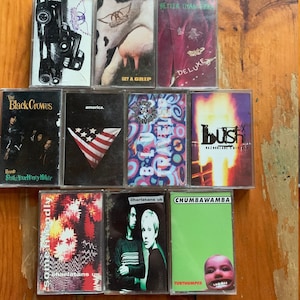 May include: A collection of ten vintage cassette tapes, including albums by The Black Crowes, Bush,  The Charlatans, and  Chumbawamba. The tapes are arranged in two rows on a wooden surface.