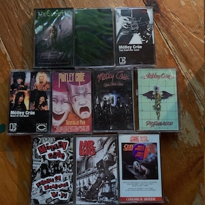 May include: A collection of vintage cassette tapes featuring rock and heavy metal bands. The tapes include albums by M&ouml;tley Cr&uuml;e, Megadeth, and Ozzy Osbourne. The album covers display band logos, album titles, and various graphics. The tapes are arranged on a wooden surface.