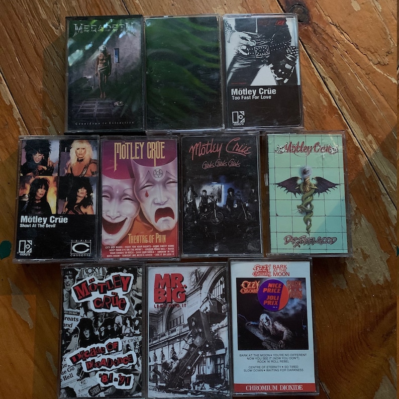 May include: A collection of vintage cassette tapes featuring rock and heavy metal bands. The tapes include albums by M&ouml;tley Cr&uuml;e, Megadeth, and Ozzy Osbourne. The album covers display band logos, album titles, and various graphics. The tapes are arranged on a wooden surface.
