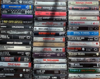 Op de afbeelding: Een verzameling vintage cassettebandjes, waaronder artiesten als The Smiths, Talking Heads en The Fixx. De bandjes zijn in rijen op een houten oppervlak gerangschikt.