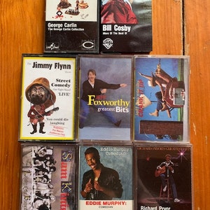 May include: A collection of eight cassette tapes featuring stand-up comedy routines by George Carlin, Bill Cosby, Jimmy Flynn, Jeff Foxworthy, Eddie Murphy, and Richard Pryor. The tapes are arranged in two rows of four, with the titles and performers' names clearly visible.