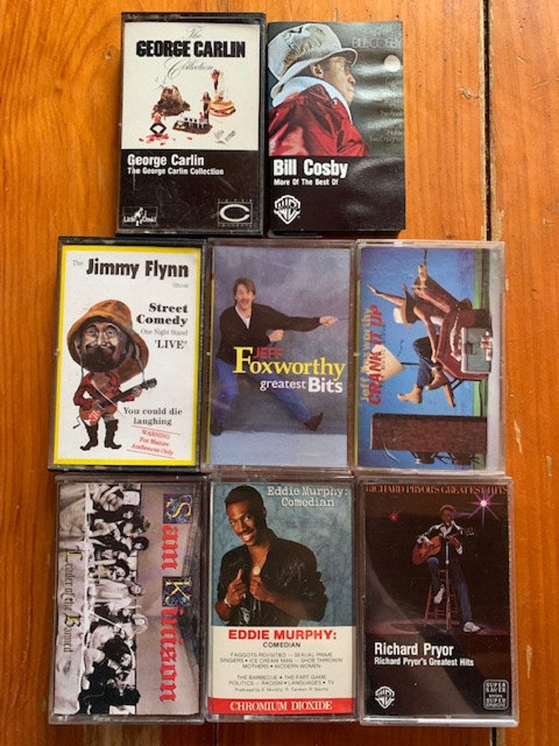 May include: A collection of eight cassette tapes featuring stand-up comedy routines by George Carlin, Bill Cosby, Jimmy Flynn, Jeff Foxworthy, Eddie Murphy, and Richard Pryor. The tapes are arranged in two rows of four, with the titles and performers' names clearly visible.