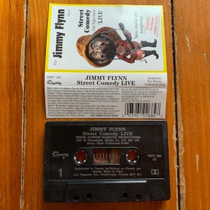 May include: A black cassette tape with a white label. The label has a picture of a man with a beard and mustache. The text on the label reads "Jimmy Flynn Street Comedy Live" and "Three Fathom Harbour Productions".