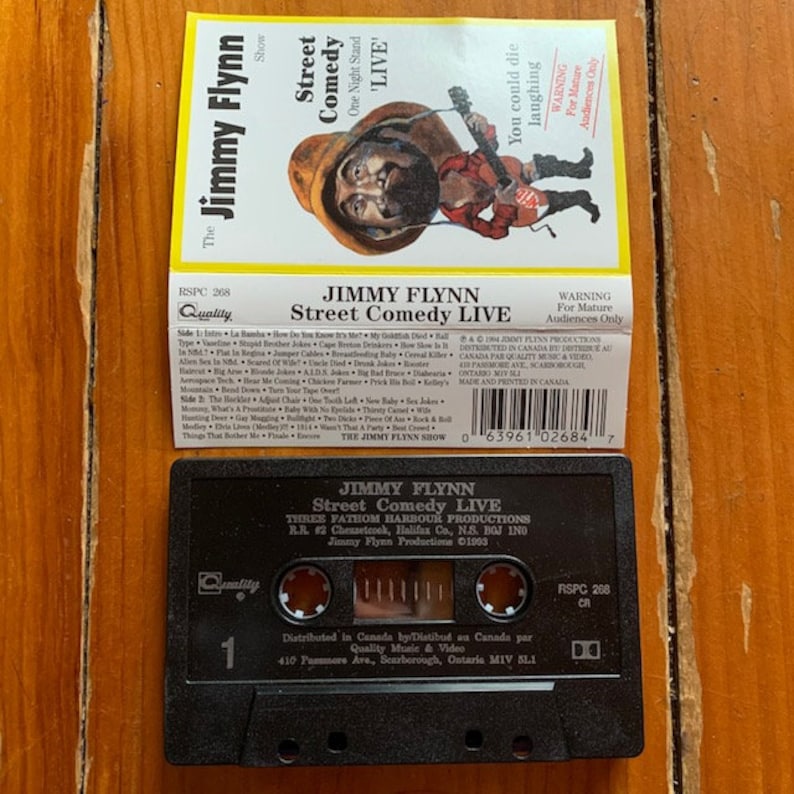 May include: A black cassette tape with a white label. The label has a picture of a man with a beard and mustache. The text on the label reads "Jimmy Flynn Street Comedy Live" and "Three Fathom Harbour Productions".