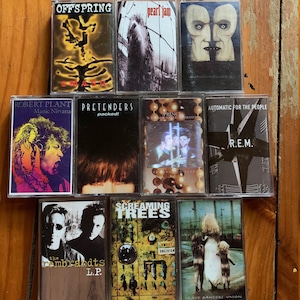 May include: A collection of nine cassette tapes featuring various artists, including Offspring, Pearl Jam, Robert Plant, Pretenders, Prince, R.E.M., Screaming Trees, and The Rembrandts. The tapes are arranged in a row on a wooden surface.