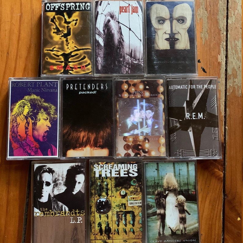May include: A collection of nine cassette tapes featuring various artists, including Offspring, Pearl Jam, Robert Plant, Pretenders, Prince, R.E.M., Screaming Trees, and The Rembrandts. The tapes are arranged in a row on a wooden surface.