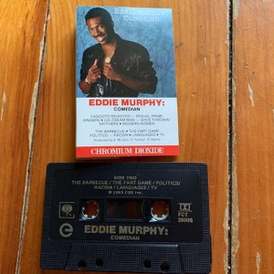 May include: A black cassette tape with a picture of Eddie Murphy on the front. The tape is labeled "Eddie Murphy: Comedian" and features the text "Side Two: The Barbecue / The Fart Game / Politics / Racism / Languages / TV" on the back. The tape is a chromium dioxide tape.