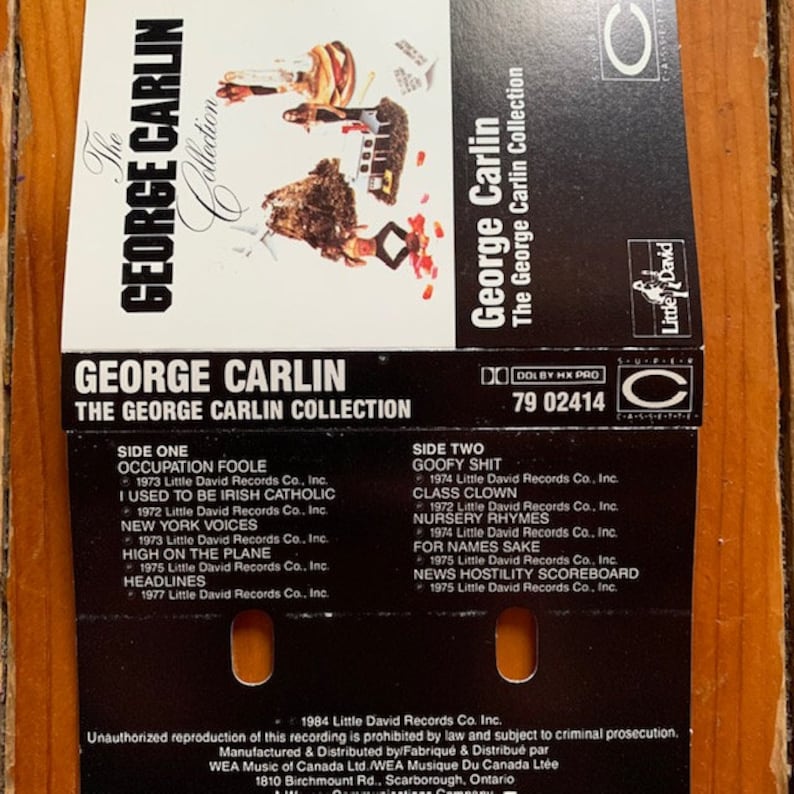 May include: A cassette tape case with a black and white photo of comedian George Carlin. The case is orange and black and has the text "The George Carlin Collection" on the front. The back of the case lists the songs on the tape, including "Occupation Foole", "I Used to Be Irish Catholic", "New York Voices", "High on the Plane", "Headlines", "Goofy Shit", "Class Clown", "Nursery Rhymes", "For Names Sake", and "News Hostility Scoreboard".