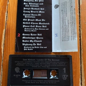 May include: A black cassette tape with a white label. The label has the title "Sam Kinison / Leader Of The Banned" and lists the songs "Detox This", "Shopping For Pets", "Sex, Videotape and Zoo Animals", "Jerry's Bastard Kid", "Lenny Bruce's Mom", "Casual Users Of Terrorism", "Old People Must Die", "Grilled Cheese Sandwich", and "Phone Call From Hell".