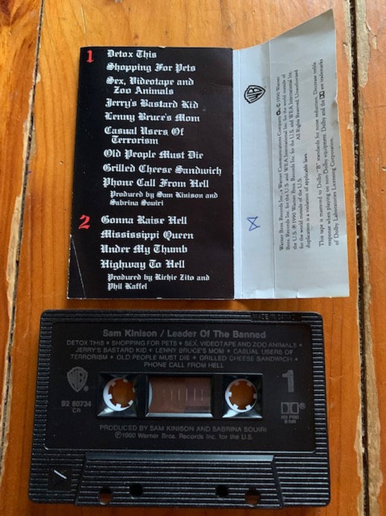 May include: A black cassette tape with a white label. The label has the title "Sam Kinison / Leader Of The Banned" and lists the songs "Detox This", "Shopping For Pets", "Sex, Videotape and Zoo Animals", "Jerry's Bastard Kid", "Lenny Bruce's Mom", "Casual Users Of Terrorism", "Old People Must Die", "Grilled Cheese Sandwich", and "Phone Call From Hell".