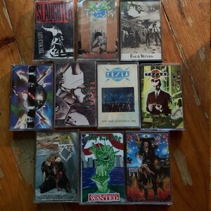 May include: A collection of vintage cassette tapes featuring various rock and metal bands. The tapes showcase album art with diverse designs, including band logos, illustrations, and text. Titles include "Slaughter", "Tesla", and "Steve Vai".