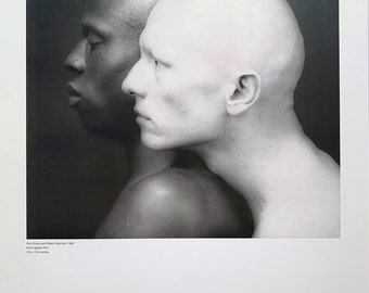 May include: Black and white photograph featuring two bald individuals in profile, one with dark skin and the other with pale skin. The image is a portrait by Robert Mapplethorpe, with text at the bottom indicating the artist's name, gallery, and dates.