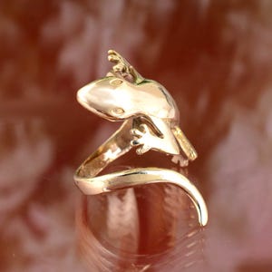 May include: A gold-toned ring featuring a detailed gecko design. The gecko is curled around the band of the ring, with its tail forming the ring's base.