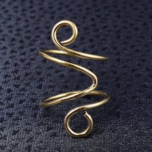 Può includere: Un anello a forma di spirale color oro. L'anello presenta un design continuo e fluido con una parte superiore e inferiore ad anello. Il metallo ha una finitura lucida, che riflette la luce su uno sfondo blu scuro. L'anello è un gioiello.