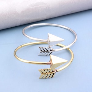 May include: Two gold and silver arrow bracelets. The bracelets are thin and delicate, with a simple arrow design. The arrowheads are triangular and the arrow shafts are adorned with a simple line pattern.